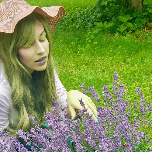 Cosplay photo of a young elvish woman with green hair and a gardening hat. Her white blouse has puffy sleeves. She is sitting in the garden behind purple flowers.